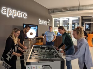 Five people are playing table football in a modern office with illuminated signs reading 